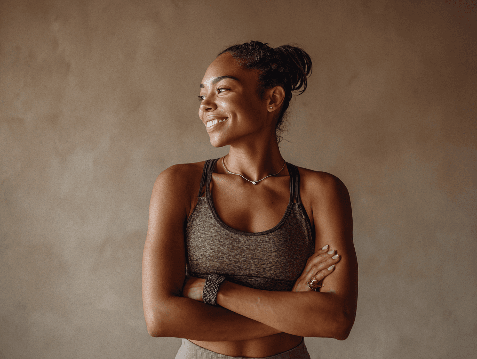 Smiling woman in athletic wear with arms crossed, looking confidently to the side against a warm neutral background